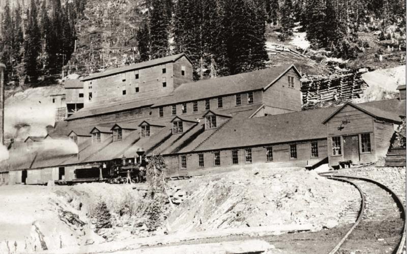 Gold King Mill at Gladstone in 1906, taken from narrow-gauge track approaching the mill, looking in northeasterly direction to front of mill and up the tram line cut beyond. Mill is in derelict state, virtually all windows broken out. Smoke is issuing from the two or three stacks visible. Locomotive No. 32 of Silverton, Gladstone &amp; Northerly Railroad stand on track in front of mill, with two unidentified crew members. San Juan County Historical Society photo