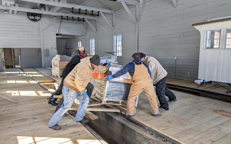 group of volunteers moves a pallet of Spacesaver shelving into storage jn the almost complete Silverton Northern Engine House. This is one of 12 pallets of materials of the units acquired for the Archinve. They will more than double the vault storage space available. As a side note, the picture also portrays the new look of the engine house interior, a welcome contrast to the dingy, poorly lit space of former days. On the right, can be seen the tail-end of the Society’s famous Casey Jones, snugly stored awa