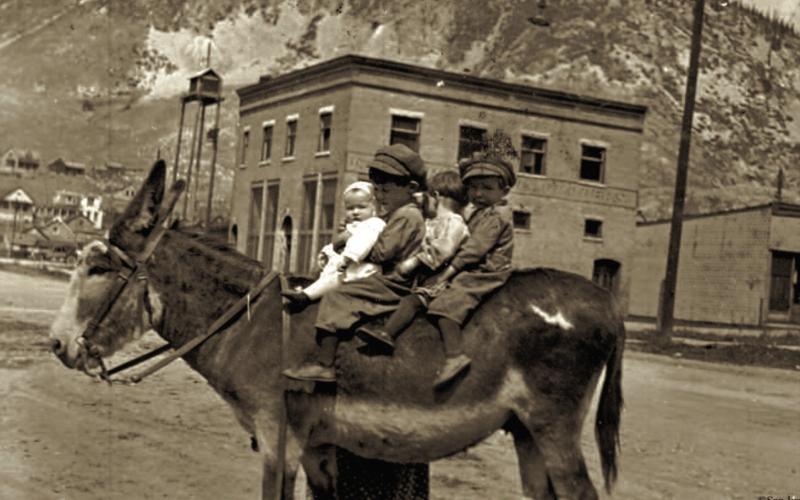 Photo courtesy of San Juan County Historical Society Four children ride a burro on 10th Street around 1920.The burro’s name was Paragron, owned by Kate Brown, mother of Nellie Jane Hill, In the background are F.O. Sherwood building (now the Avon Hotel) and the old fire siren tower on the alley between Greene and Blair. Note the original cornice on the Avon building (not replaced after a fire in the 1930s).