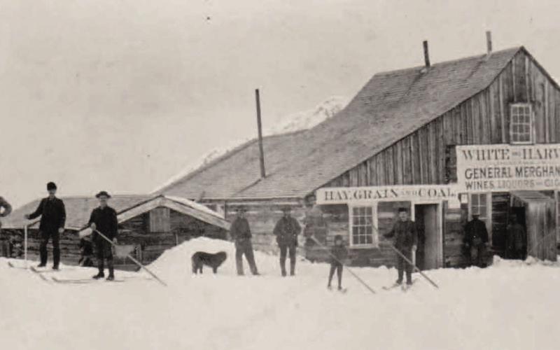 The White &amp; Harvey General Store at Mineral Point. Its signs advertise wine, liquor, cigars, hay, grain, coal, hardware, tinware, and groceries. Thirteen people, nine on skis, are shown, along with two large dogs. The view is looking north. Photo Courtesy of Silverton Mountain.