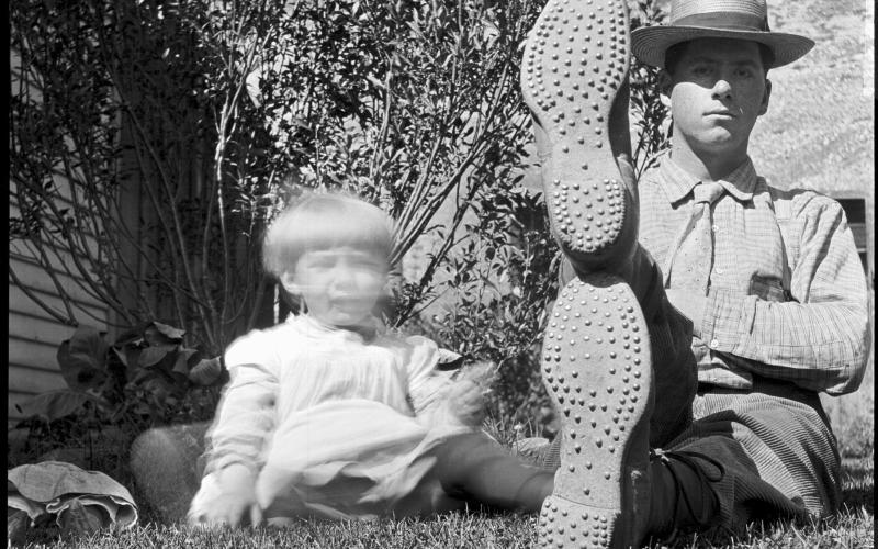 Edgar James Dyson sitting in yard of the Dyson home with one foot propped atop the other. Displays the cleated soles and heels of his hiking boots. Entitled 'Engineering Feat' by the photographer. The blurred subject at left is young Dan McNaughton. Image Courtesy of San Juan Historical Society