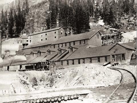 Gold King Mill at Gladstone in 1906, taken from narrow-gauge track approaching the mill, looking in northeasterly direction to front of mill and up the tram line cut beyond. Mill is in derelict state, virtually all windows broken out. Smoke is issuing from the two or three stacks visible. Locomotive No. 32 of Silverton, Gladstone &amp; Northerly Railroad stand on track in front of mill, with two unidentified crew members. San Juan County Historical Society photo