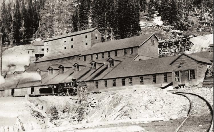 Gold King Mill at Gladstone in 1906, taken from narrow-gauge track approaching the mill, looking in northeasterly direction to front of mill and up the tram line cut beyond. Mill is in derelict state, virtually all windows broken out. Smoke is issuing from the two or three stacks visible. Locomotive No. 32 of Silverton, Gladstone &amp; Northerly Railroad stand on track in front of mill, with two unidentified crew members. San Juan County Historical Society photo